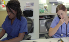 Two medical professionals sit at a desk in an office while one (right) talks on the phone