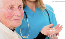 An elderly woman with pills in her hand stands in front of a nurse