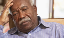 An elderly man sits looking down with a hand to his head